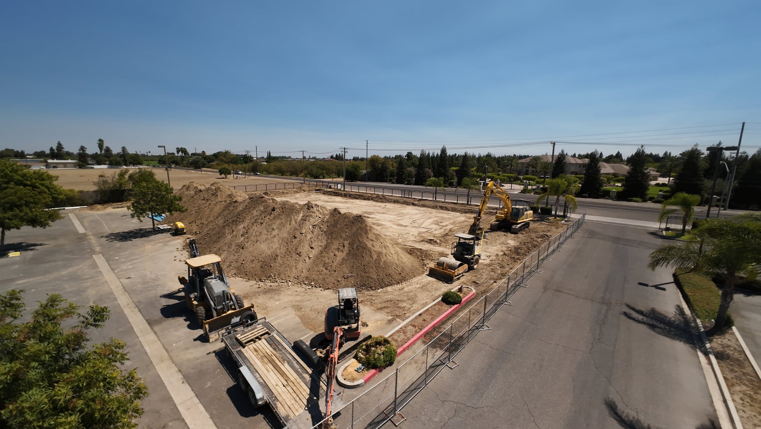 Aerial view of the fenced church site with soil piles, a yellow excavator, and a roller on the graded pad