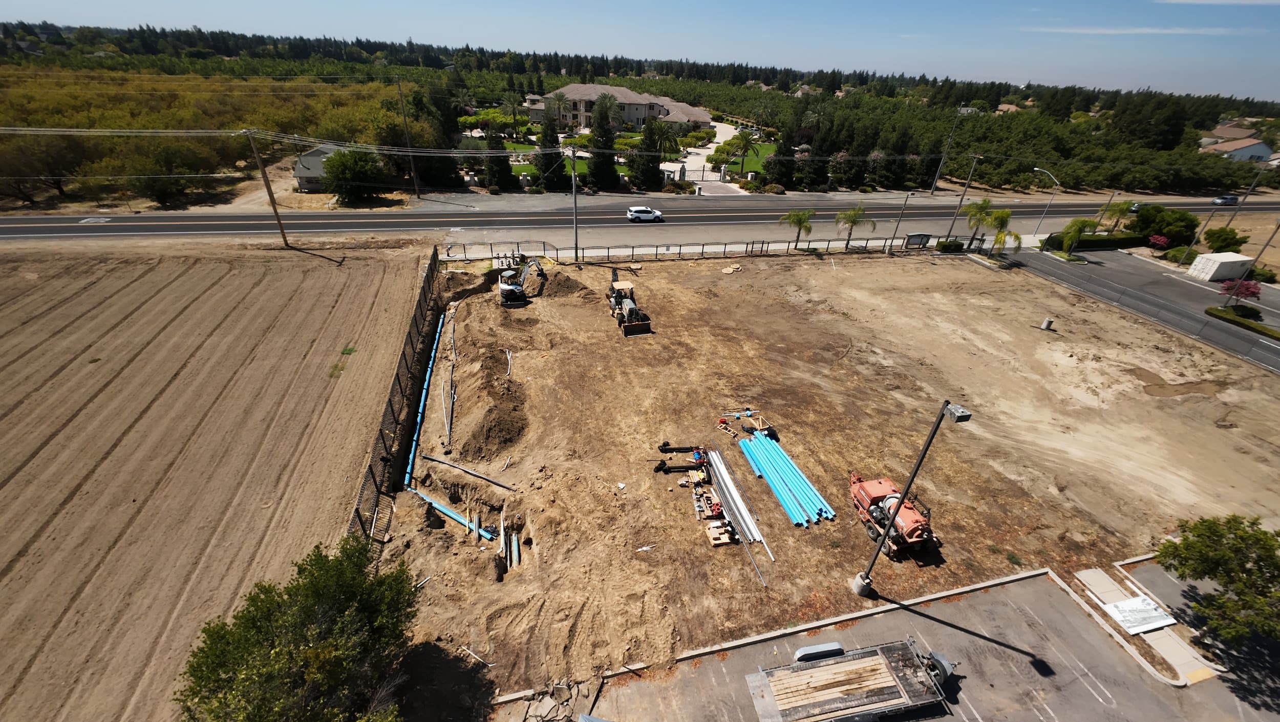 Aerial view of the cleared church site with a trench, stacked blue water pipe, and construction equipment